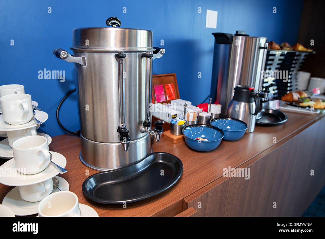 Großer Edelstahl-Kaffeespender, der auf einem Holzbuffet im Frühstücksbereich eines Hotels aufgestellt ist, mit weißen Keramikbechern und Schüsseln. Stockfoto