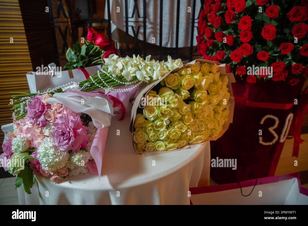 Elegante Geschenke, die mit Perlen verziert sind, vorbereitet für ein besonderes Jubiläumsereignis. Verschiedene Luxusblumensträuße inklusive weißer und rosafarbener Rosen Stockfoto