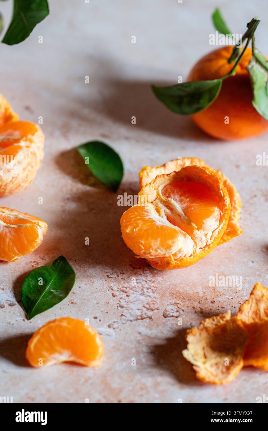 Frische Mandarinen mit geschälten Segmenten, strukturierter Schale und glänzenden grünen Blättern, die auf einer rustikalen hellen Oberfläche angeordnet sind, präsentieren eine lebendige Zitrusfrucht Stockfoto