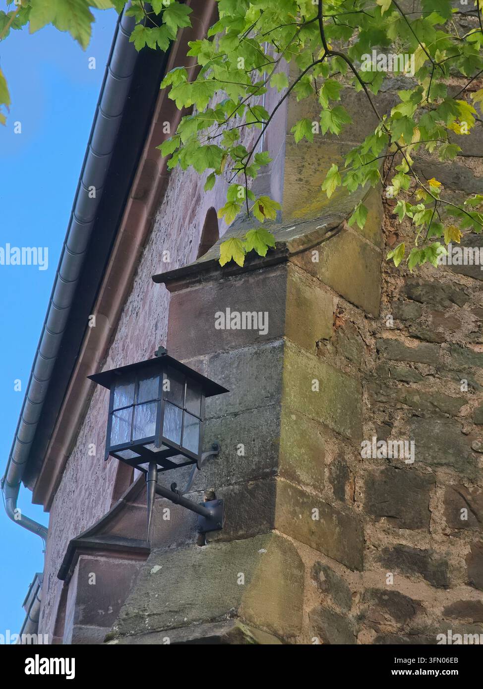 Antike Wandlaterne, die an einer historischen Mauer aus Stein und Schiefer montiert ist, mit grünen Ästen unter einem blauen Himmel. - Smartphone-aufgenommenes Stockfoto