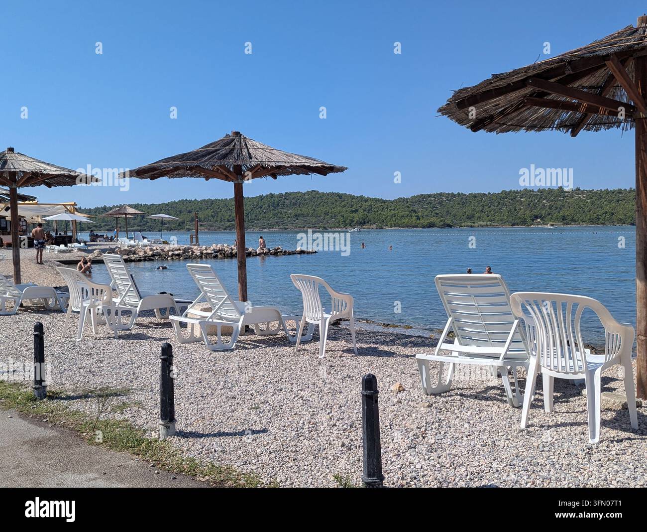 Pirovac, Kroatien, 16. August 2026 Leere weiße Loungestühle und Strohsonnenbeschattungen säumen einen mit Kies bedeckten Uferstreifen neben ruhigem blauem Wasser, mit Strand, Meeresufer, Ufer Stockfoto