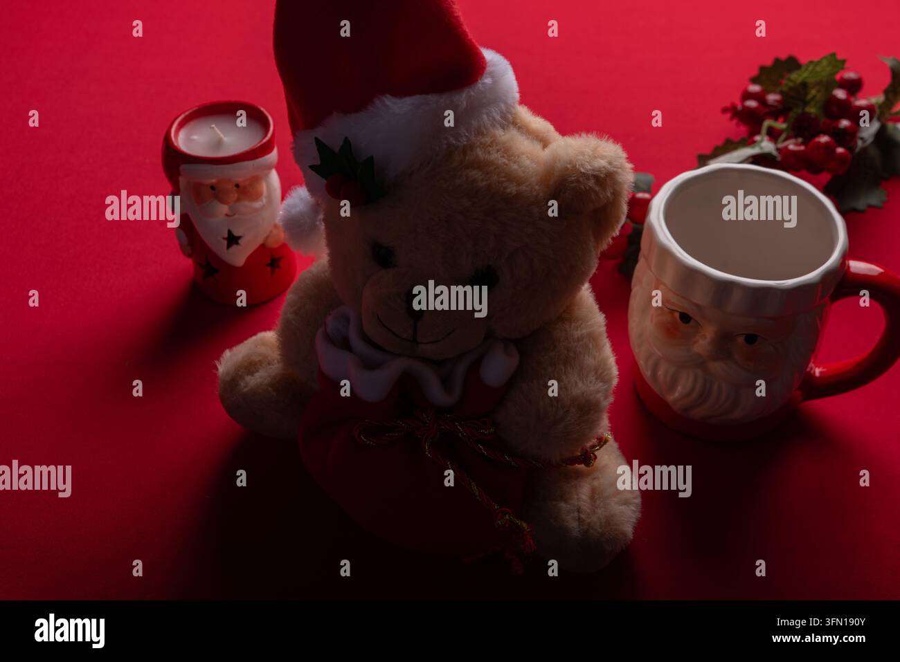 Weiches Plüschbären mit Weihnachtsmütze und festlichen Dekorationen, darunter Weihnachtsbecher und Kerze, auf einer lebhaften roten Oberfläche. Warme Studiolichtverhältnisse betonen den Plüsch Stockfoto