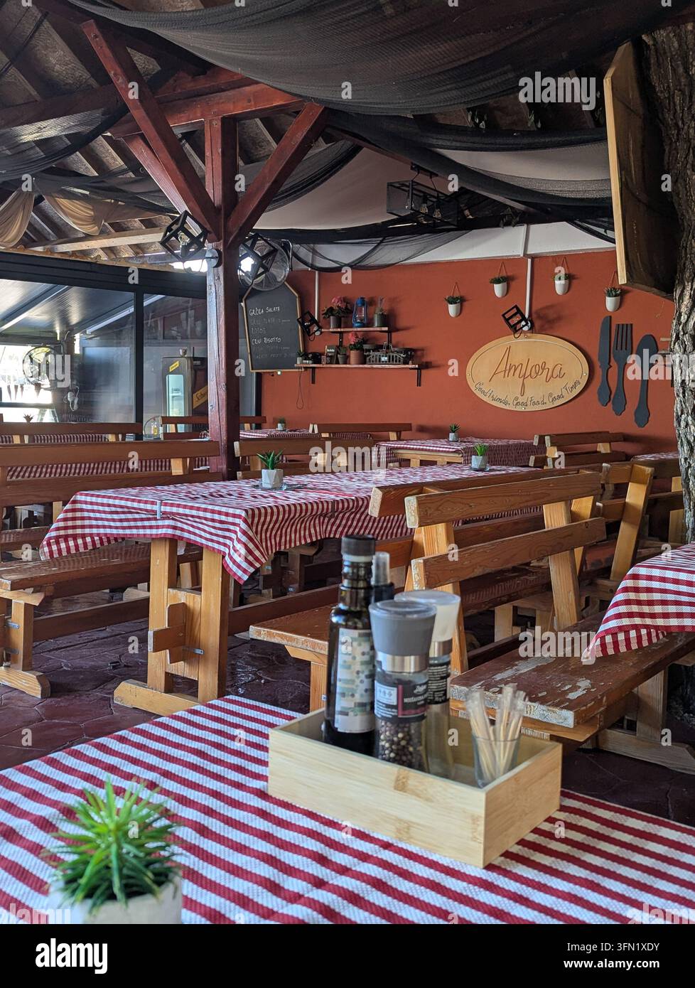 Pirovac, Kroatien, 16. August 2026 Rustikales Open-Air-Restaurant-Interieur mit Holzbänken, karierten Tischdecken, hängender Dekoration, Terracotta-Wand Stockfoto