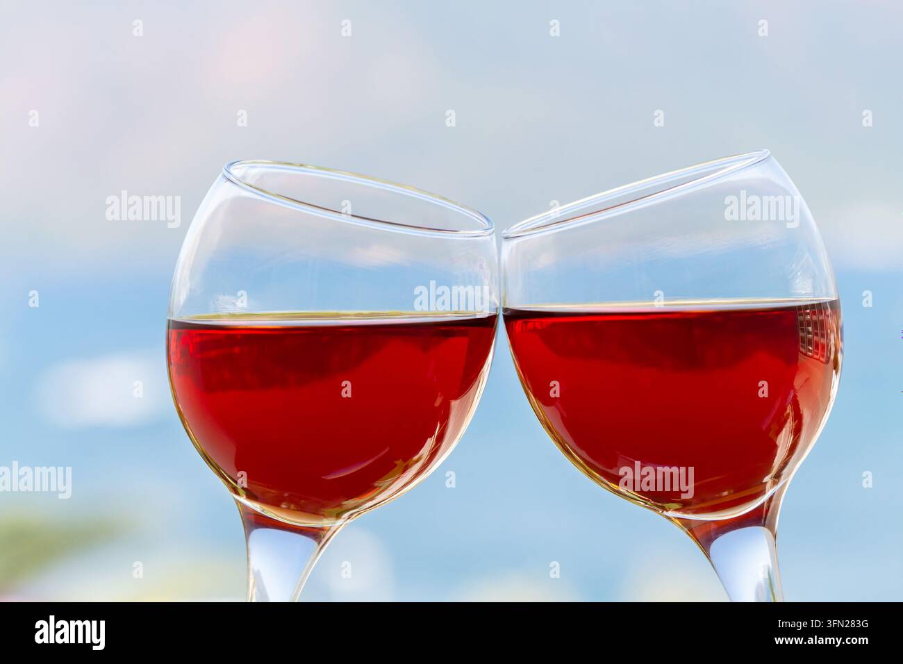 Nahaufnahme von zwei Stielgläsern mit Roséwein, die sich bei einem Toast aneinanderlehnen, vor einem sanft unscharfen blauen Hintergrund, der eine entspannte Außenumgebung suggeriert Stockfoto