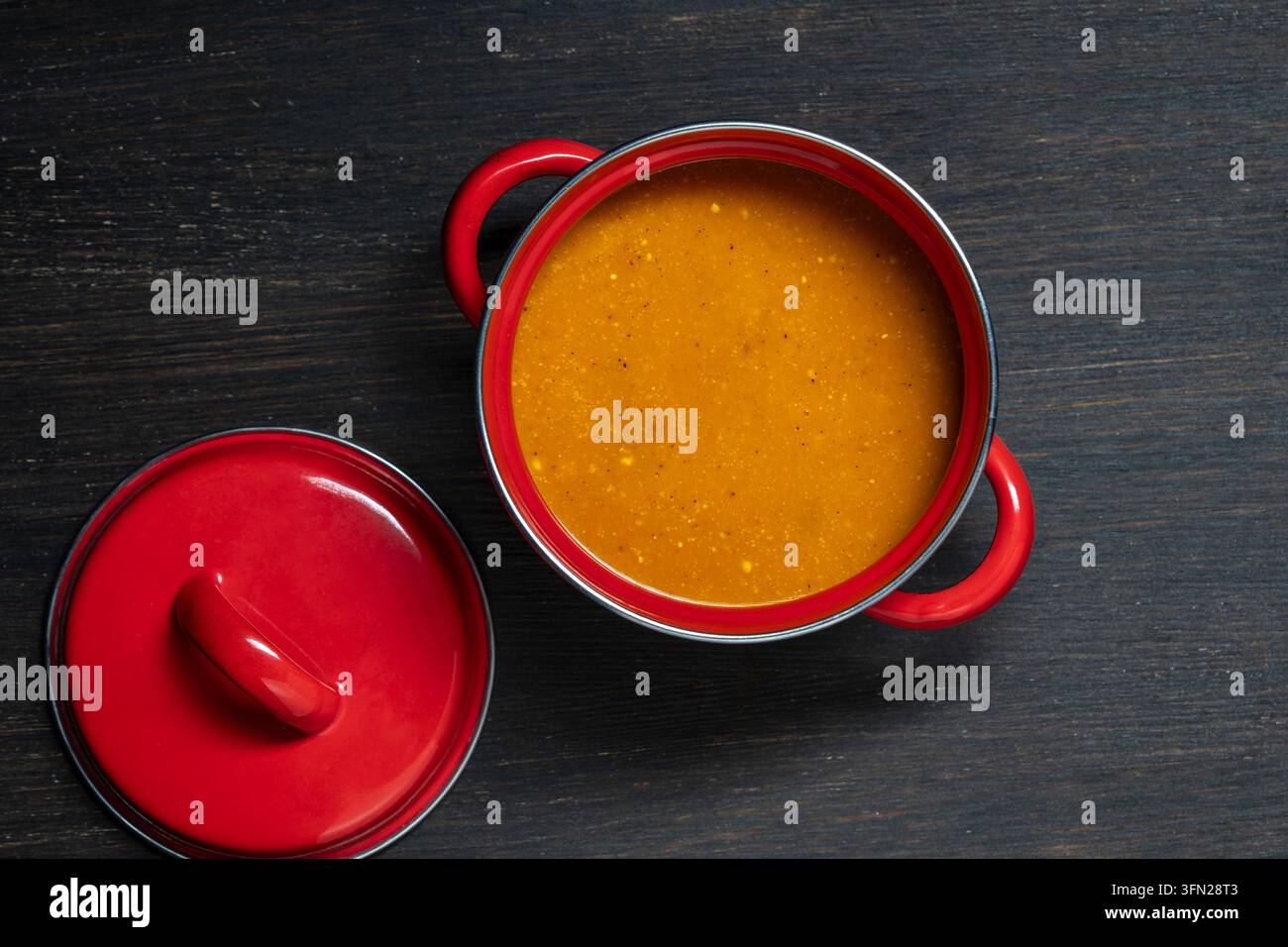Hausgemachte Kürbissuppe in einem roten Topf. Oberansicht der köstlichen heißen Orangencremesuppe in einem lebendigen roten Emailtopf mit offener Deckel, serviert auf rustikalem Holzta Stockfoto