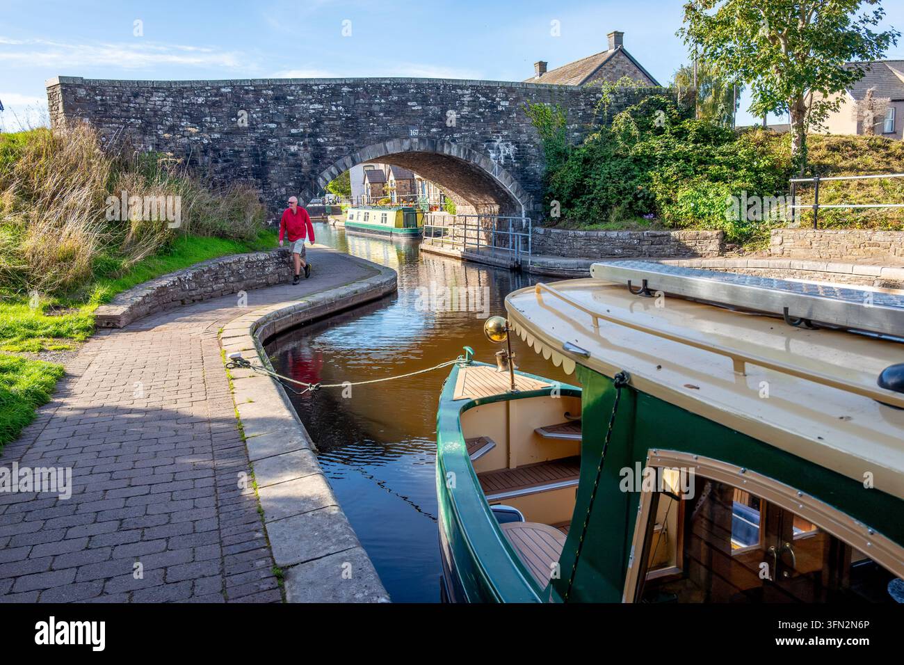 Ein Mann geht am Kanalufer entlang in der Nähe einer historischen Steinbogenbrücke, während ein grünes Kanalfahrzeug im ruhigen Wasser an einem sonnigen Nachmittag ruht Stockfoto