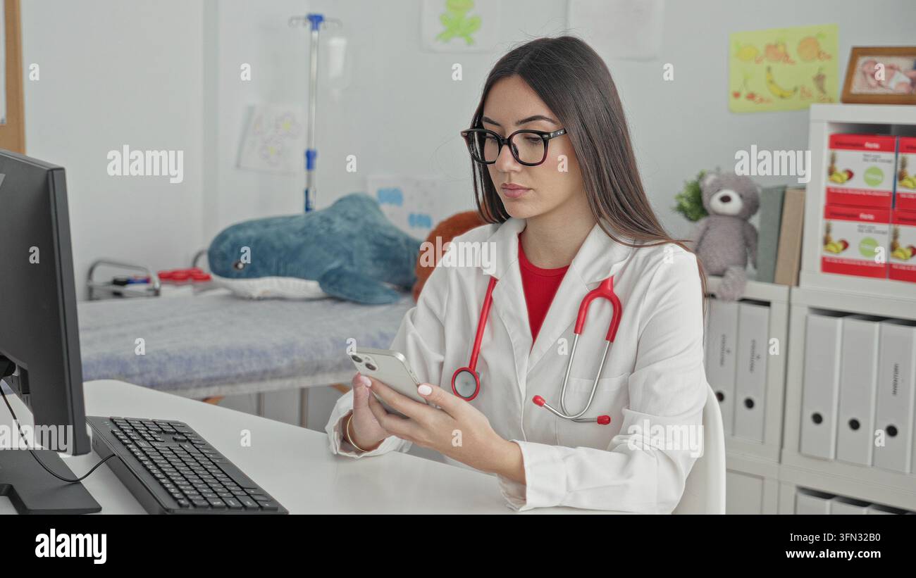 Junge Ärztin mit Stethoskop, die Smartphone hält, Hände sichtbar und scrollen, während sie am Computerpult in einem Gebäude sitzt; ruhige Konzentration. Stockfoto