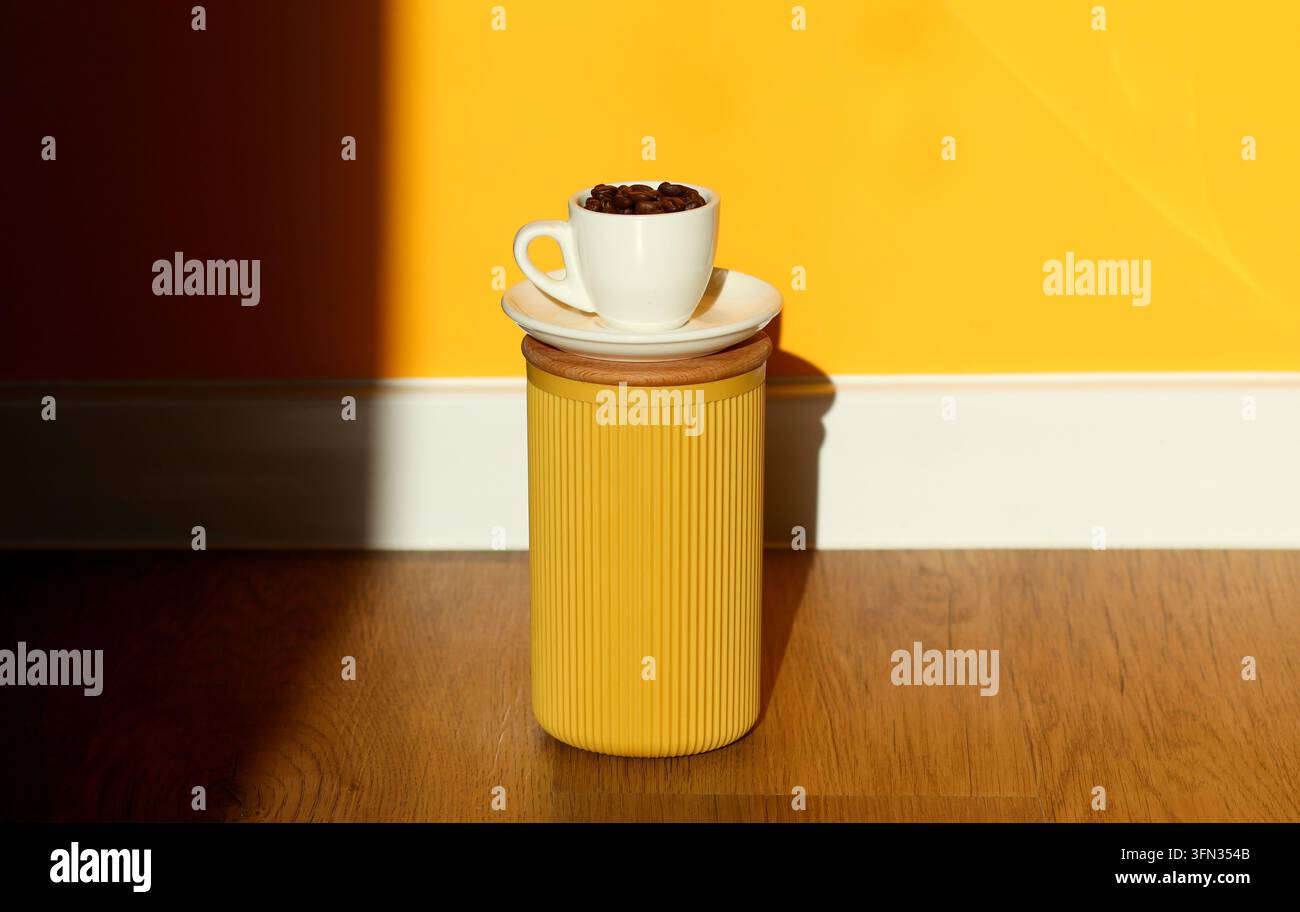 Eine weiße Tasse gefüllt mit gerösteten Kaffeebohnen auf einer Untertasse auf einem gelben zylindrischen Behälter vor einem lebendigen gelb-schwarzen Wandhintergrund. Minima Stockfoto