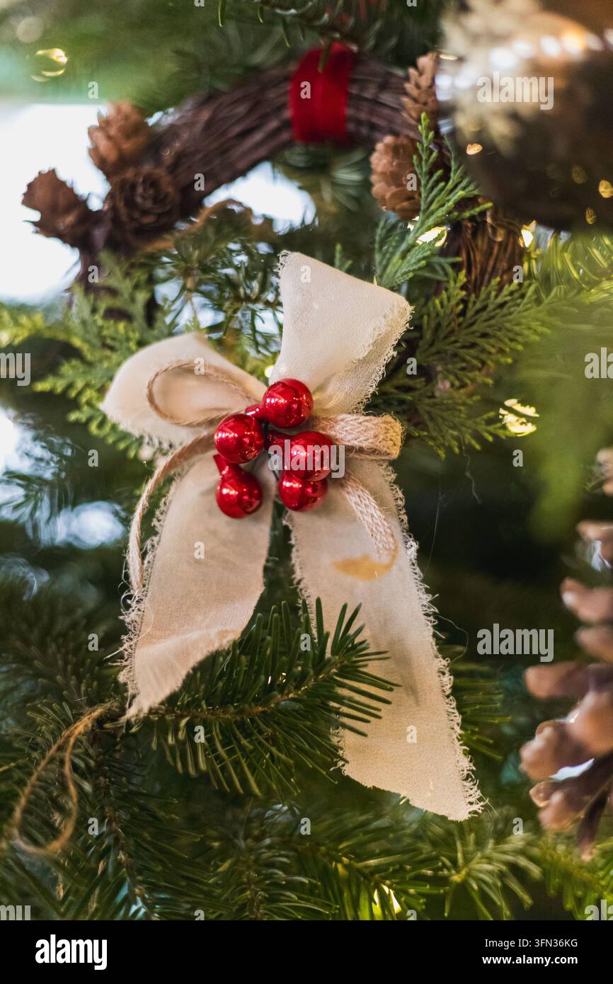 Nahaufnahme eines rustikalen handgefertigten Weihnachtsschmucks mit einer natürlichen Sackleinen-Schleife, roten Beeren und Schnur, der zwischen immergrünen Zweigen und Kiefernzapfen hängt Stockfoto