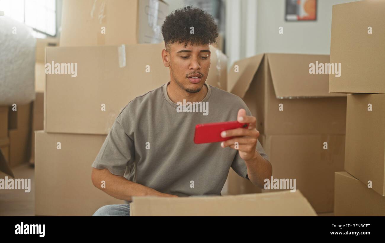 Mann hält rotes Smartphone und gestikuliert mit der Hand zwischen Kartonumzugskisten in einem Gebäude; Ablenkung am Packtag. Stockfoto