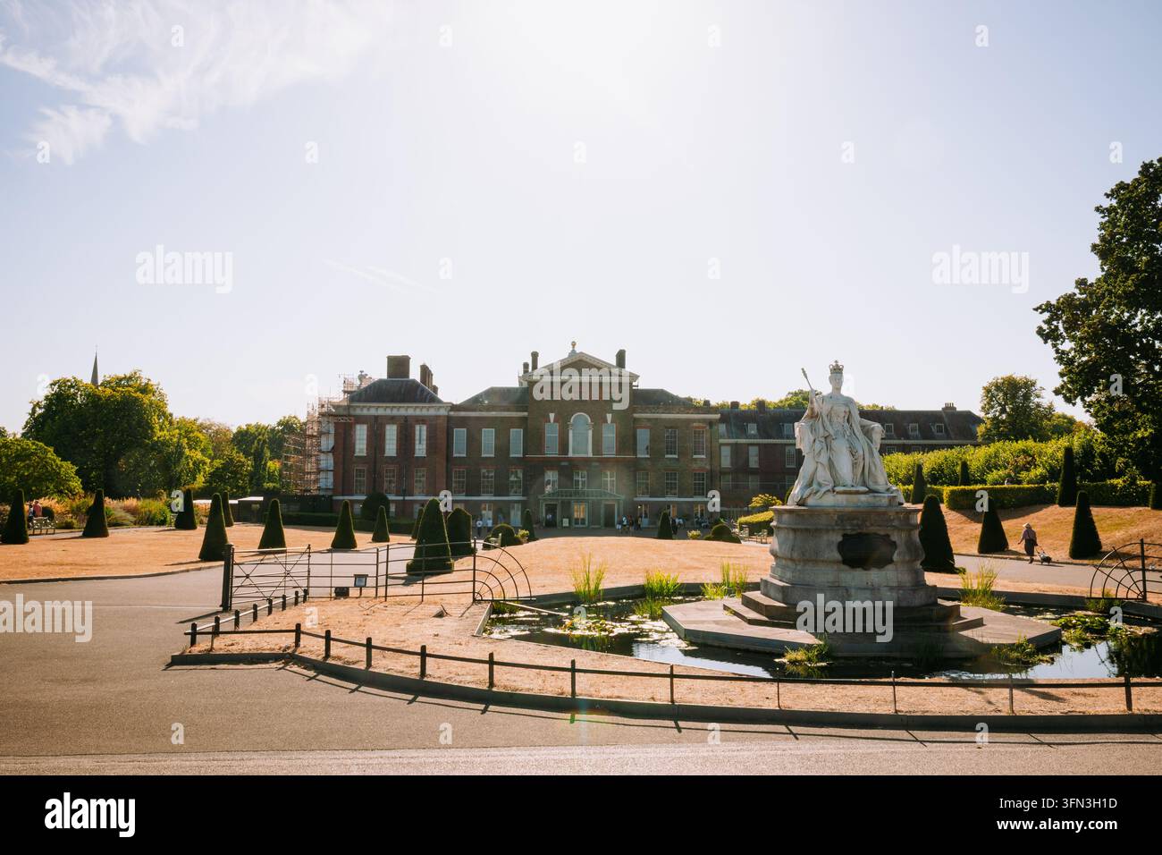 London, England, 12. August 2026. Weite Aufnahme der Backsteinfassade von Kensington Palace mit der Statue von Königin Victoria und einem Teich im Vordergrund unter hellem Sommerlicht Stockfoto