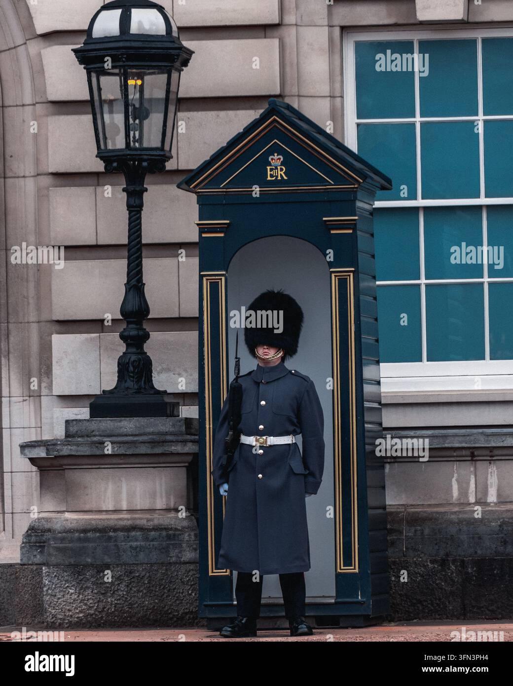 London, UK - 28. März 2021: Ein Queen's Guard in einem Bärenfellhut und einem Umhang steht an einer Wachkabine vor dem Buckingham Palace. Stockfoto