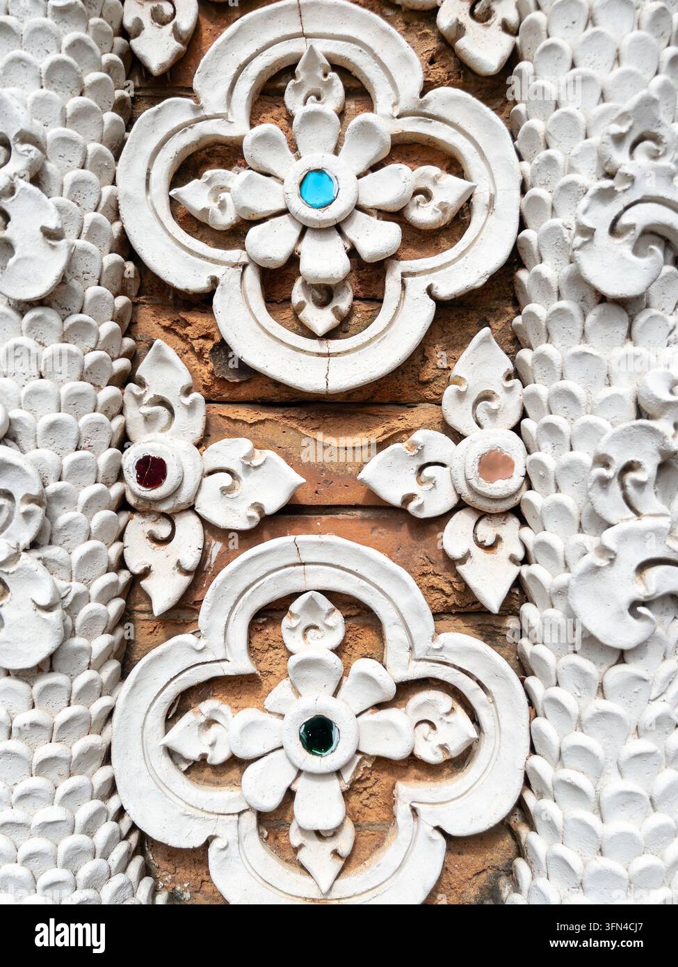 Beschreibung: Nahaufnahme eines kunstvollen weißen Stuckblumenornaments mit Glasmosaikdetails an einer alten Tempelmauer in Thailand. Stockfoto
