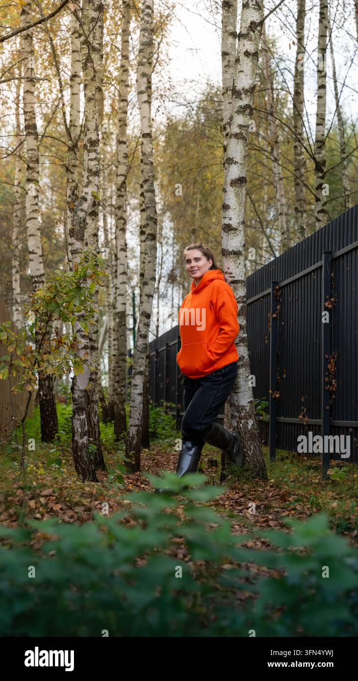 Junge Frau in orangefarbenem Kapuzenpullover und Gummistiefeln genießt frische Herbstluft neben einer Birke und einem Metallzaun in einem ruhigen Hain Stockfoto