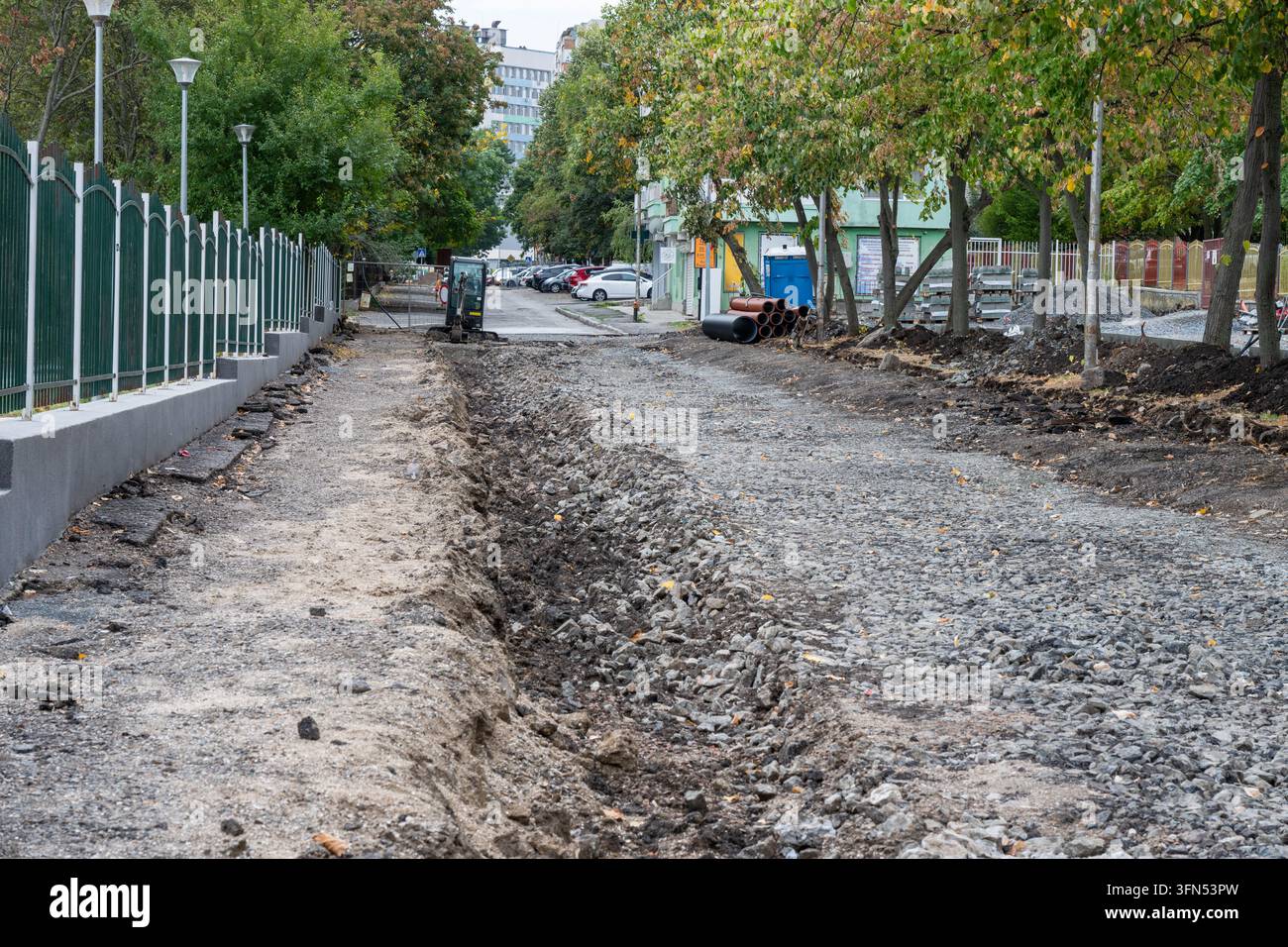 Eine zerrissene städtische Straße und ein ausgehobener Bürgersteiggraben, gefüllt mit losem Schotter, verlaufen parallel zu einem grünen Metallzaun und einer Baumreihe während der Kommunal Stockfoto
