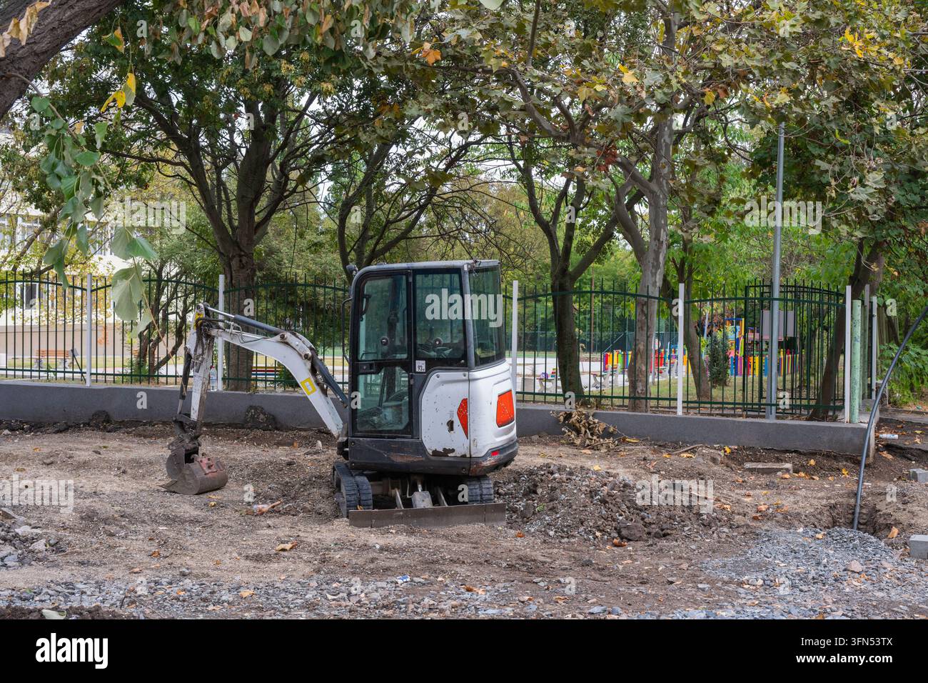 ein weißer Minibagger, der auf einer Schotter- und Kiesbaustelle geparkt ist, umgeben von Bäumen und einem grünen Metallzaun. Stockfoto