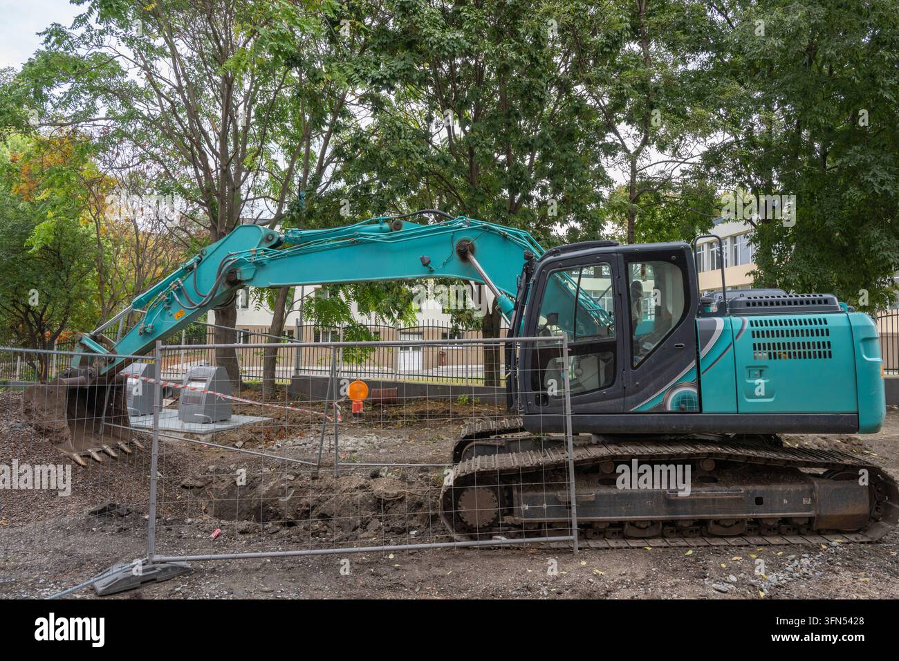 ein blaugrüner Kettenbagger, der hinter einem provisorischen Metall-Sicherheitszaun auf einer Schotter- und Schmutz-Baustelle mit Bäumen und einem Gebäude im Hintergrund geparkt ist Stockfoto