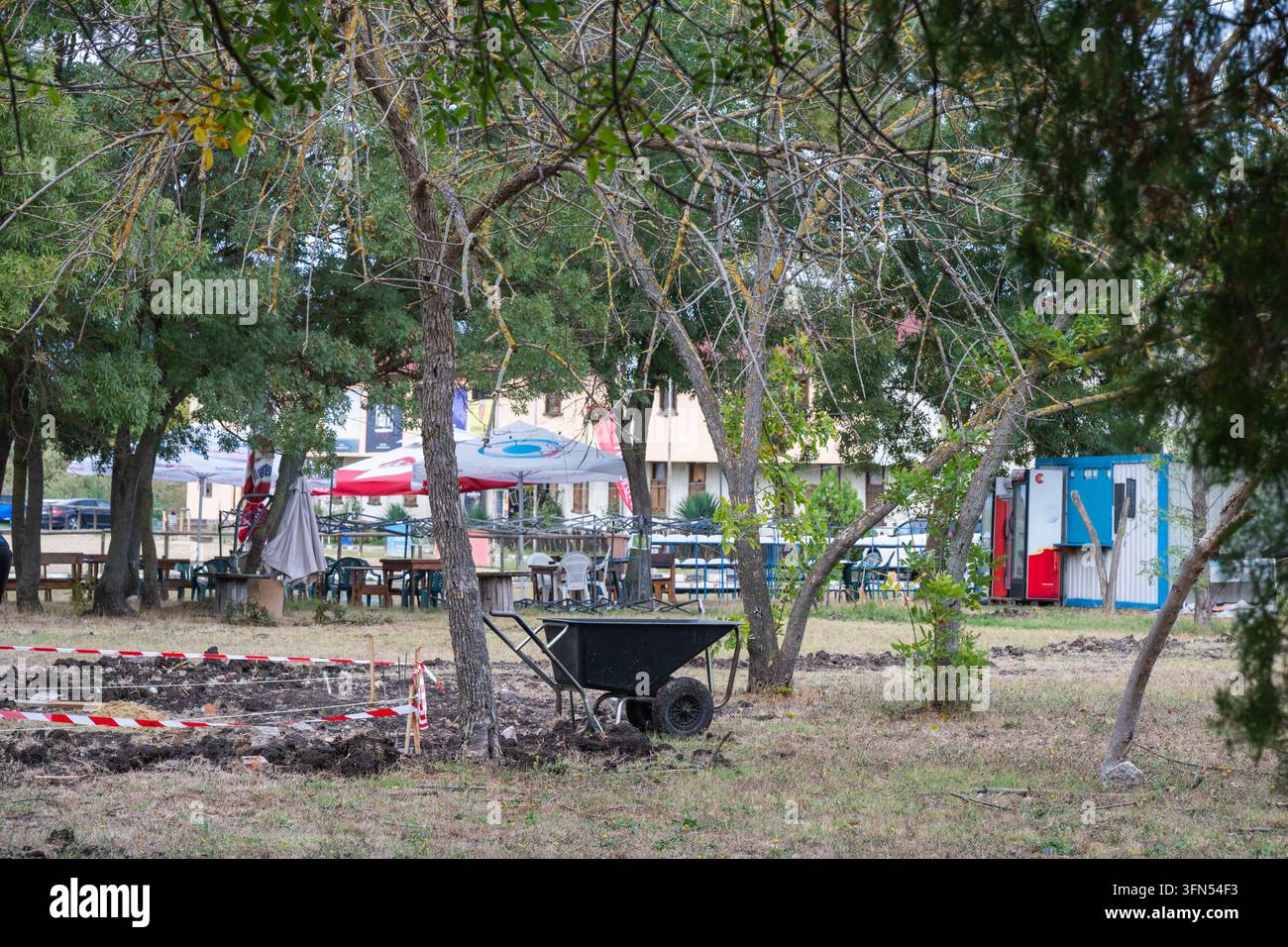 Eine schwarze Schubkarre steht neben einem abgesperrten Landschaftsbereich in einem bewaldeten Außenpark in der Nähe einer Freizeitanlage und schattiger Patiotische Tische. Stockfoto