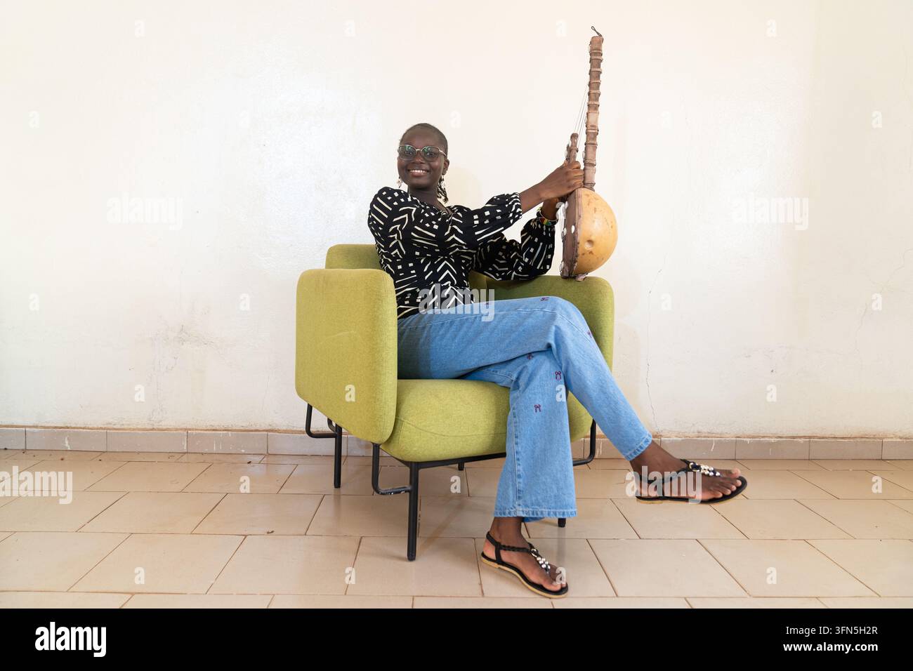Lächelnde afrikanische Frau hält ein traditionelles westafrikanisches Saiteninstrument in Innenräumen und feiert Musik, kulturelles Erbe, weibliche Kunstfertigkeit und Erhaltung Stockfoto