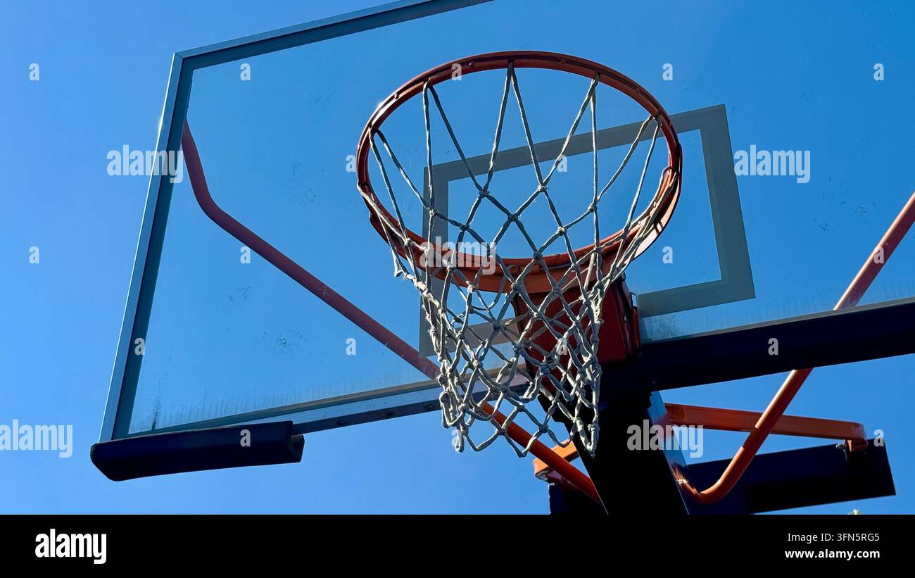 Seitenansicht eines Outdoor-Basketballkorbs mit Glasrückwand, orangefarbenem Ring und Netz vor einem hellblauen Himmel. Stockfoto