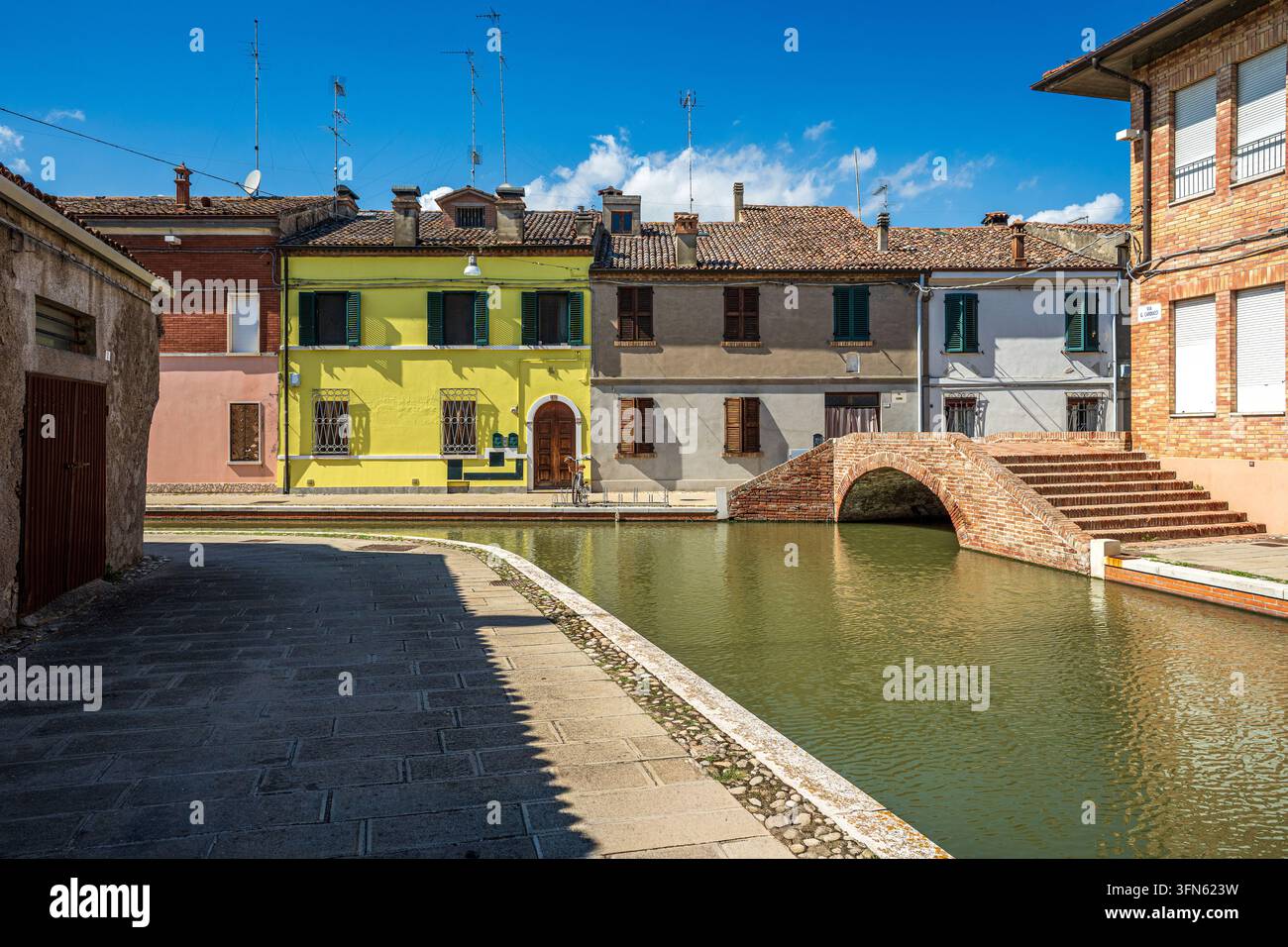 Charakteristische Ansicht von Comacchio mit einem ruhigen Wasserkanal, der entlang historischer Reihenhäuser verläuft, darunter eine auffällige gelbe Fassade. Rechts ein s Stockfoto