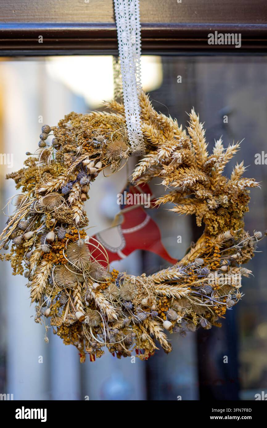 Handgefertigter rustikaler Kranz aus getrocknetem Weizen und Wildblumen, der an einem Fenster hängt, natürliche saisonale Dekoration mit kunstvollen botanischen Texturen. Stockfoto