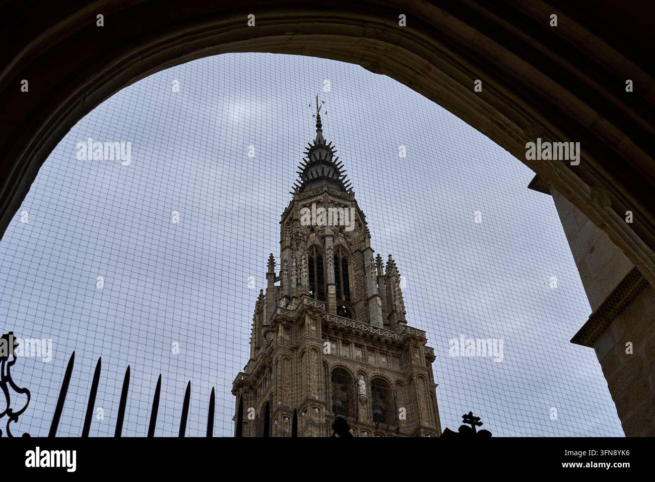 Glockenturm der Kathedrale von Toledo, eingerahmt von einem dunklen Steinbogen und eisernen Geländern unter einem grauen Himmel, Spanien Stockfoto