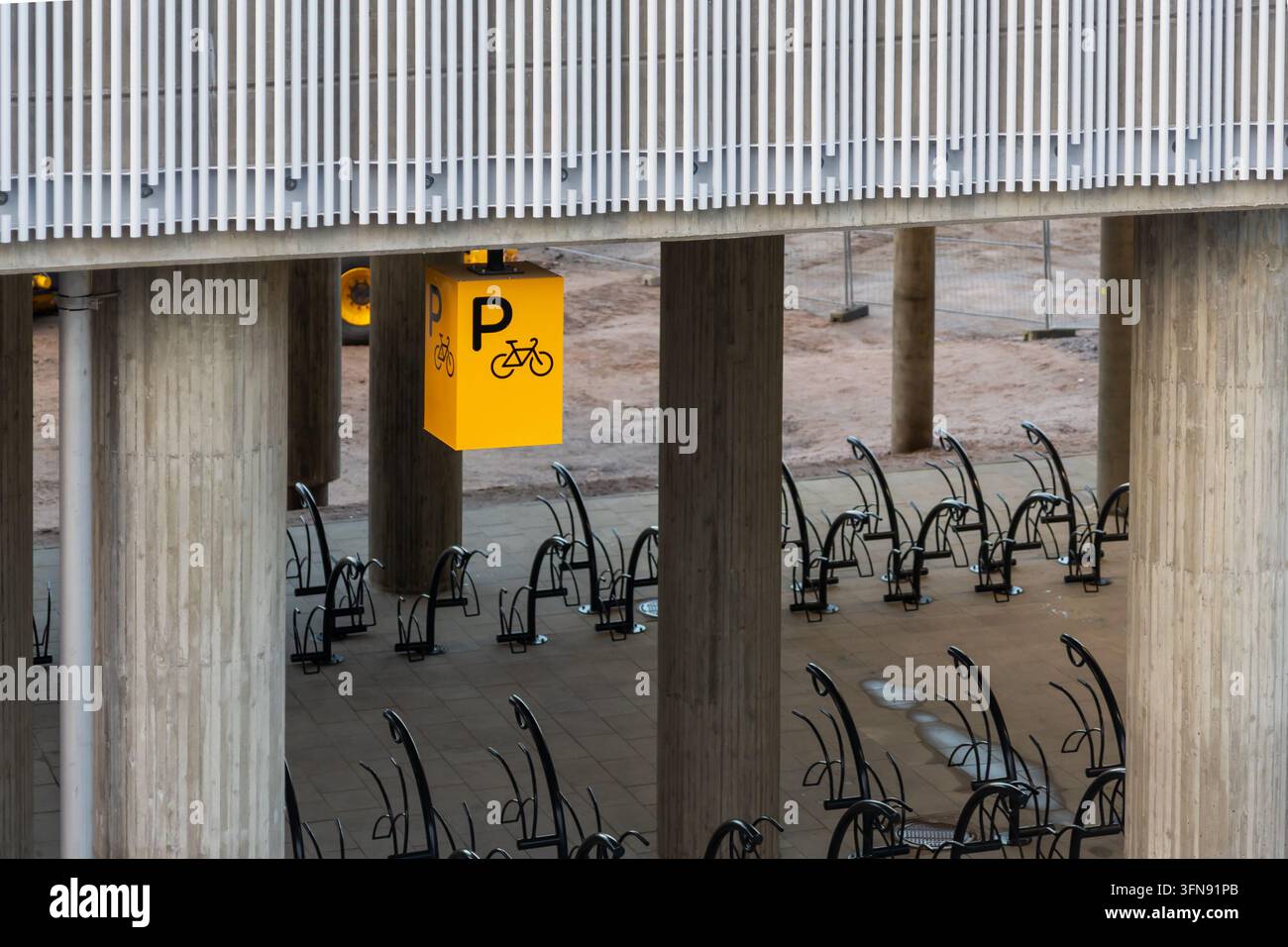 Espoo, Finnland - 1. Oktober 2026: Ein gelbes Fahrradparkzeichen, das unter einer Betonbrücke und einem Bauwerk mit schwarzen Fahrradständern hängt, die installiert sind auf Stockfoto
