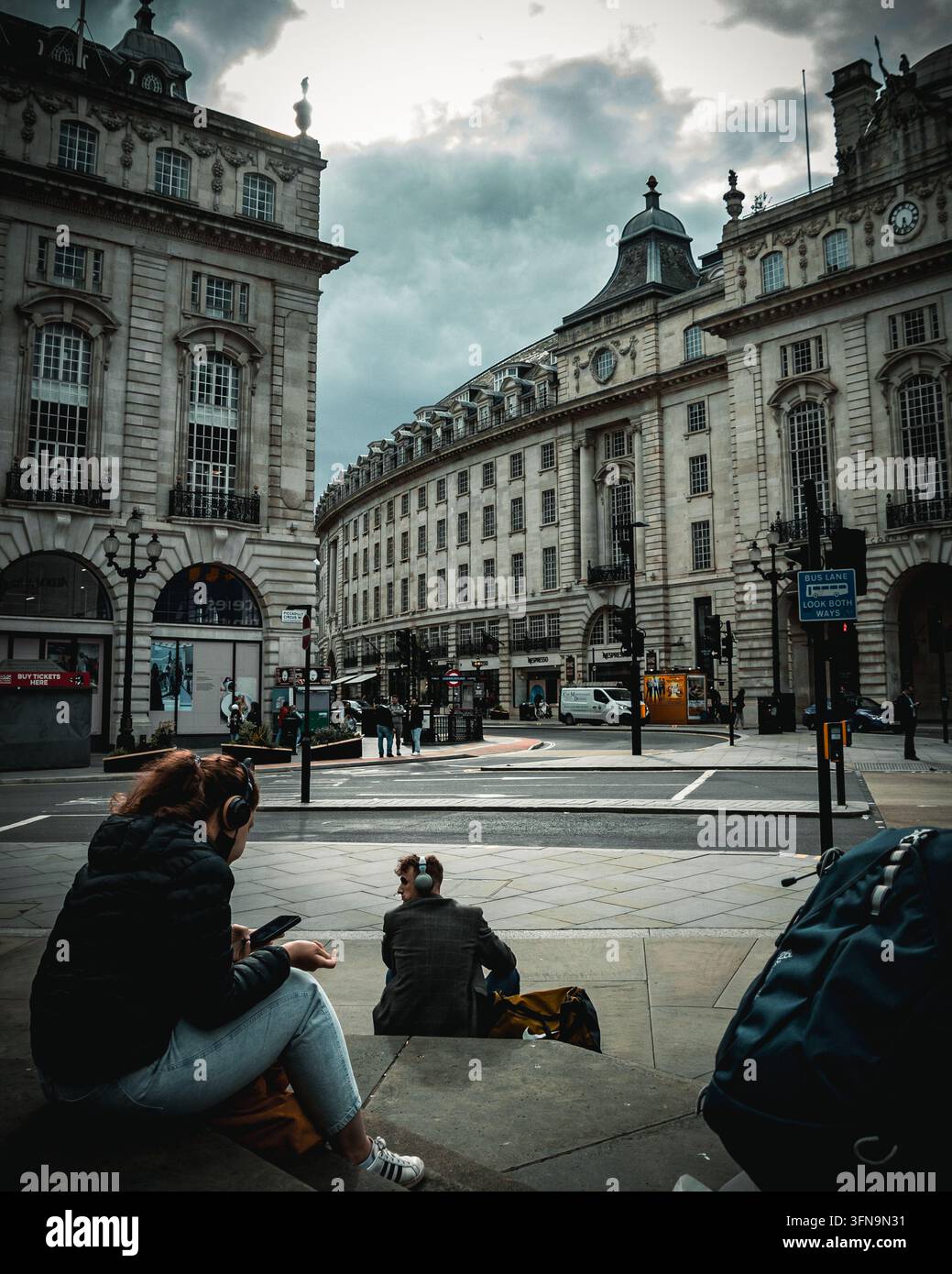 Piccadilly Circus, London, UK - 22. Juni 2021: Zwei Personen mit Kopfhörern sitzen auf Steinstufen mit Blick auf die geschwungenen Fassaden des Regent Street Quadrant Stockfoto