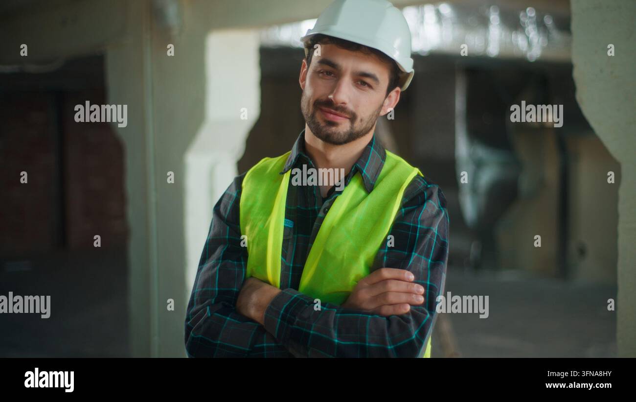 Fröhlicher kaukasischer Bauarbeiter lächelt in die Kamera auf der Baustelle, zufriedener, selbstbewusster männlicher Arbeiter posiert als Bauunternehmer mit Helm, Ingenieur Stockfoto
