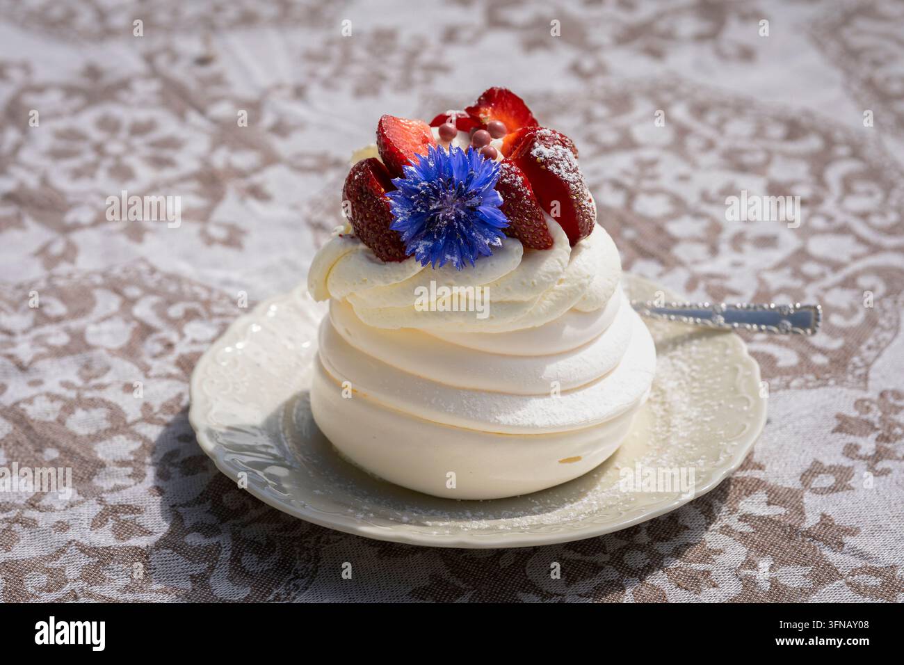 Pavlova-Dessert mit Schlagsahne, frischen Erdbeeren und einer leuchtenden blauen Kornblume garniert, mit Puderzucker bestäubt und auf einem eleganten Teller serviert. Stockfoto