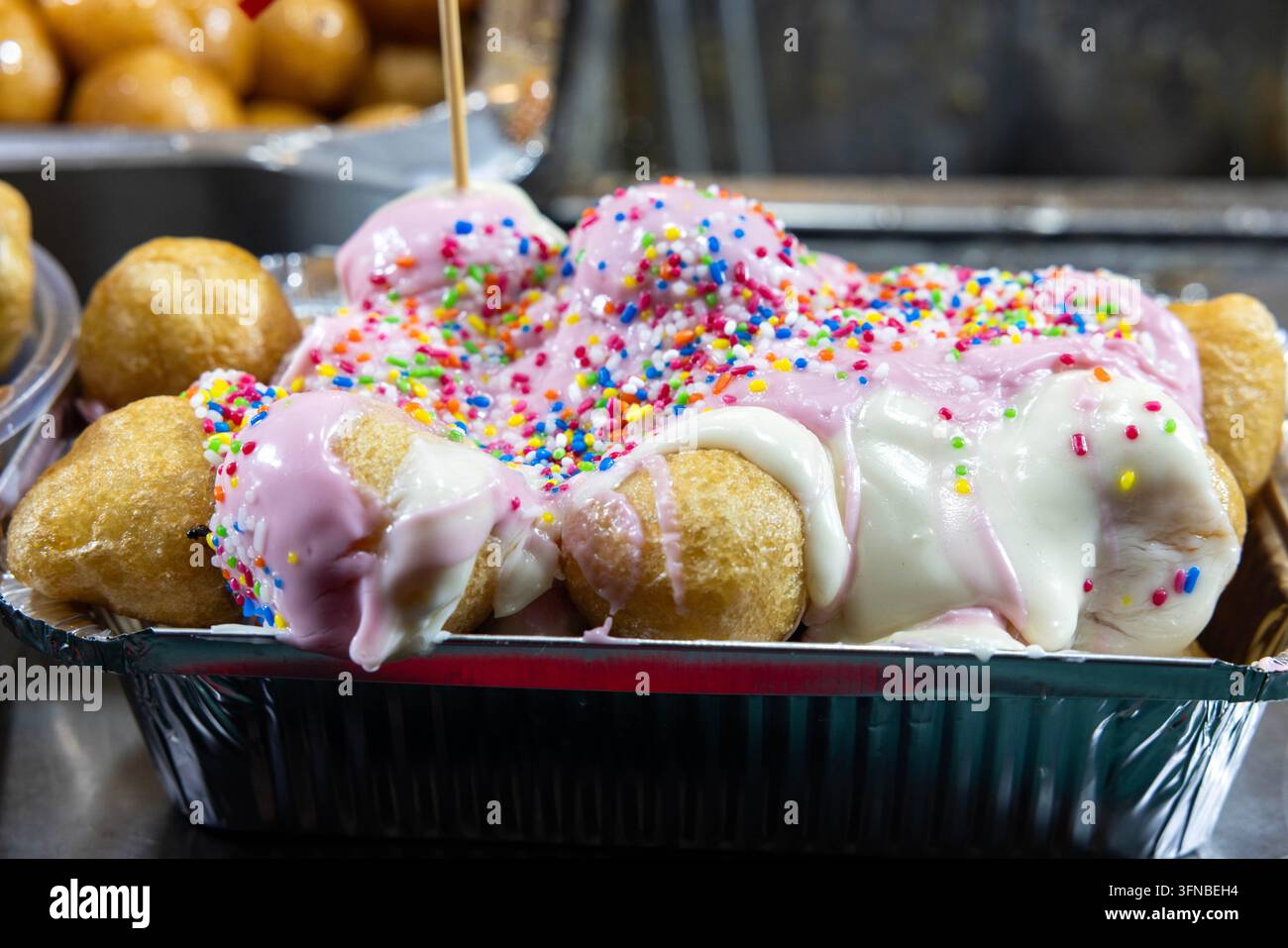 Frisch zubereitete griechische Loukoumades mit weißer Schokolade, Bananenscheiben, rosa Sahne und bunten Streuseln, serviert auf einem Weihnachtsmarkt während Stockfoto