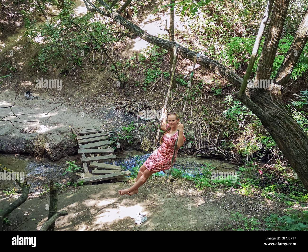 Lächelnde Frau schwingt auf einem Bungee-Seilschaukel über einen natürlichen Süßwasserbach in der Nähe der Myrne-Quelle in der Ukraine. Aktive Outdoor-Erholung und Lifestyle-Porträt - Smartphone-aufgenommenes Stockfoto