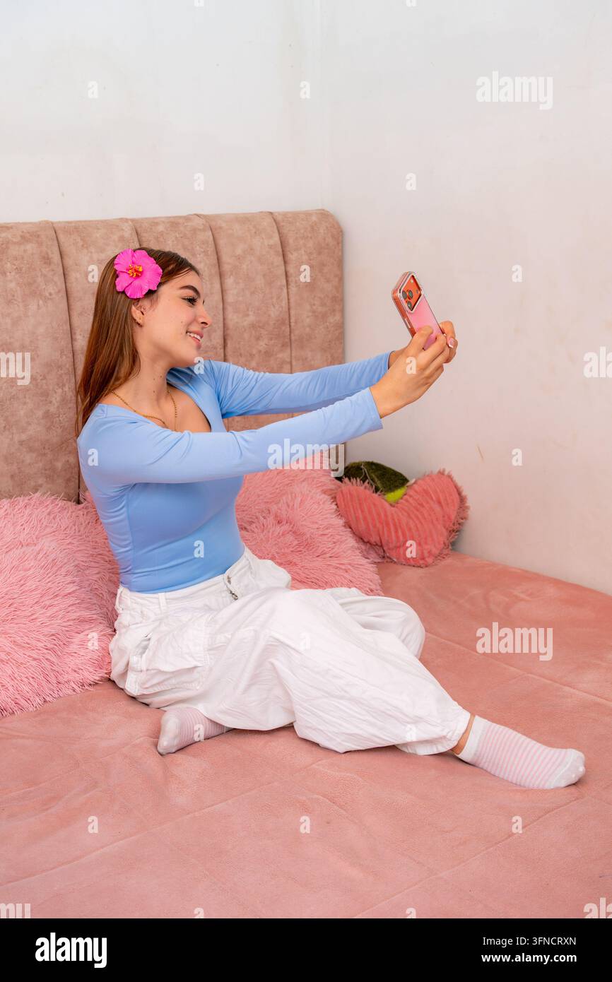 Junge erwachsene Frau lächelt und entspannt sich auf einem gemütlichen rosa Bett, während sie mit ihrem Smartphone ein Selfie macht und einen Moment der Freude und Muße einfängt, in he Stockfoto