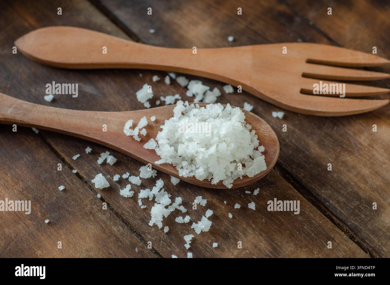 Grobes Salz ruht in einem Holzlöffel. Ein Holzgabel liegt neben dem Löffel. Eine staubige Holzoberfläche verleiht rustikalen Charme. Natürliches Licht hebt das Salz hervor Stockfoto