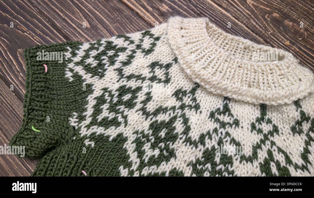 Ein weiß-grünes, handgestricktes Rundkragen mit einem Jacquardmuster liegt auf einem dunklen Holztisch neben dekorativen Blumen. Detail eines zukünftigen Pullovers. Stockfoto