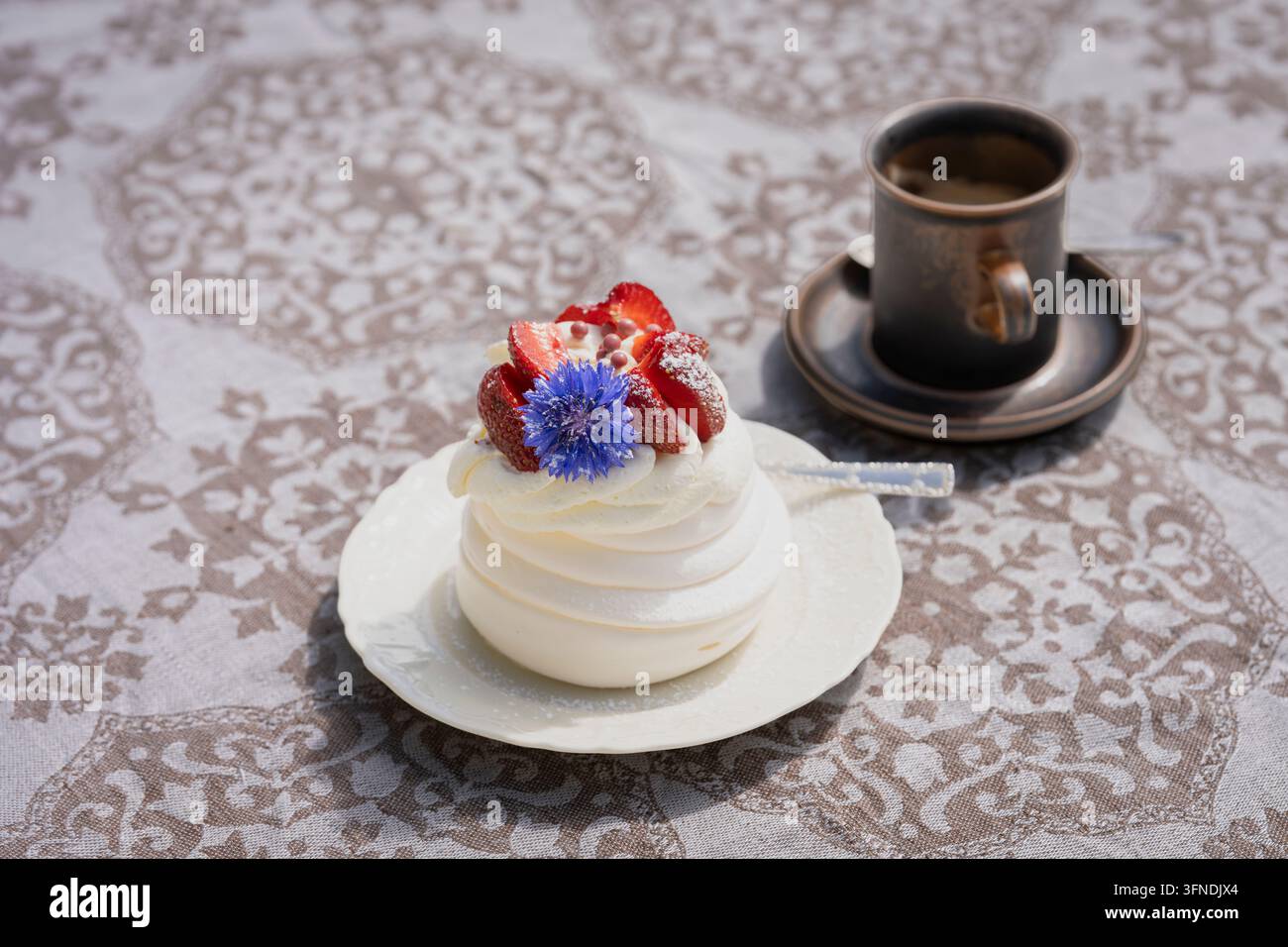 Pavlova-Dessert mit Schlagsahne, frischen Erdbeeren und einer lebhaften blauen Kornblume, serviert auf einem eleganten weißen Teller mit einer Tasse Kaffee. Stockfoto