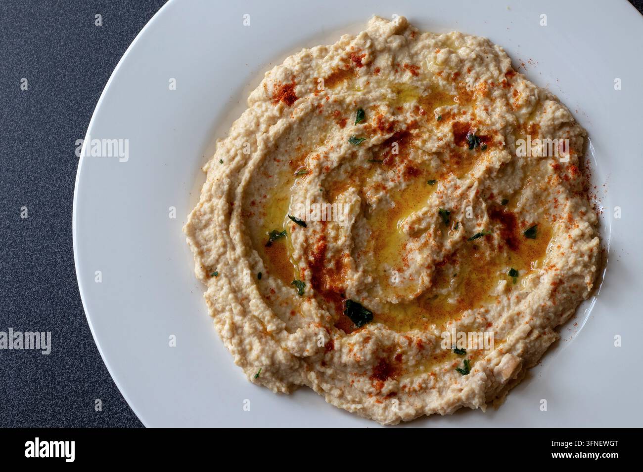 Oberansicht von cremigem Hummus, der mit Olivenöl, Paprika und gehackter Petersilie verziert ist, auf einem weißen Teller mit breitem Rand neben einer dunklen Arbeitsplatte, nah dran Stockfoto