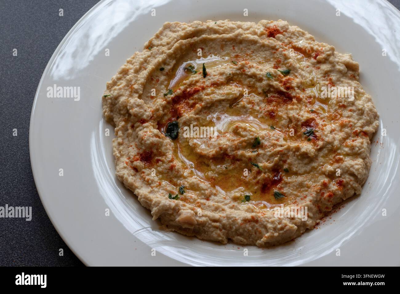 Hochwinkelansicht von cremigem Hummus mit Olivenöl, Paprika und Petersilie auf einem glänzenden cremefarbenen Teller in stimmungsvoller Tiefenbeleuchtung auf einer dunklen gesprenkelten Arbeitsplatte Stockfoto