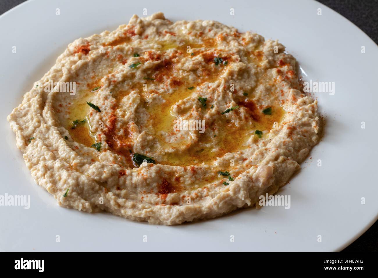 Helle 45-Grad-Ansicht von cremigem Hummus, der mit Olivenöl, Paprika und Petersilie verziert ist, auf einem großen weißen Teller mit einer dunklen Eckenplatte Stockfoto
