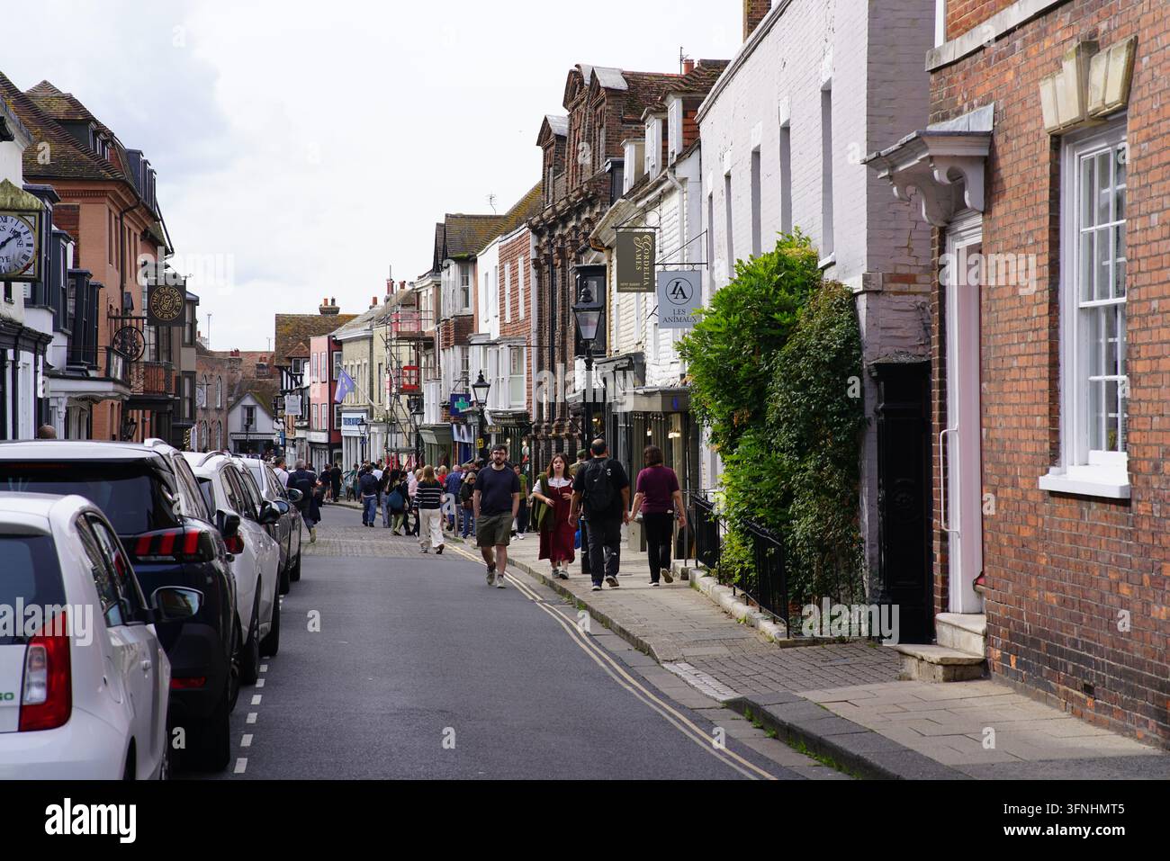 Menschen gehen auf dem Bürgersteig der High Street in Rye, zwischen den Schaufenstern und einer Backsteinmauer. Rye, England Stockfoto
