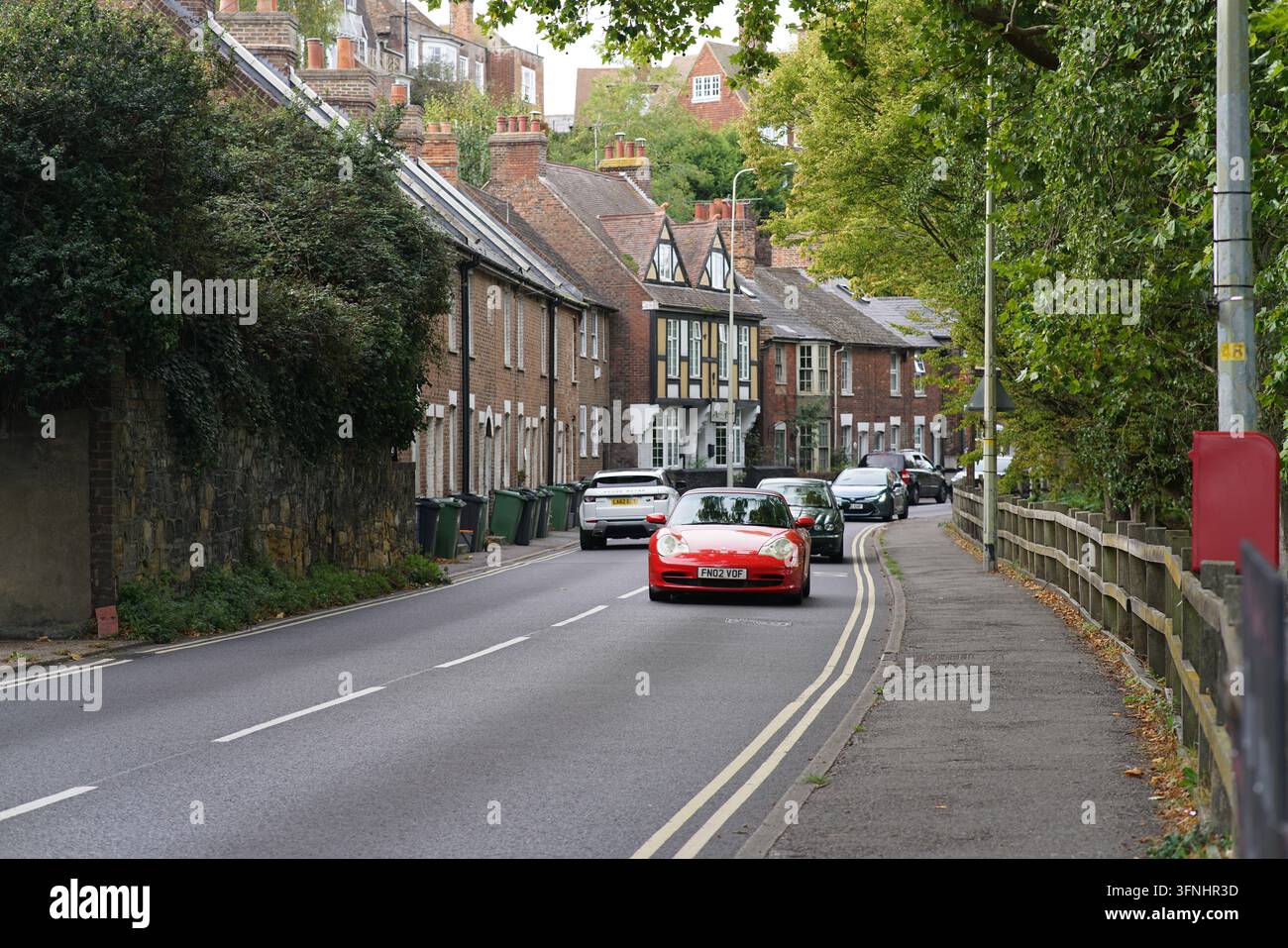 Ein rotes Auto folgt den doppelten weißen Linien auf einer Straße in Rye, zwischen einer Steinmauer und einem Bürgersteig unter Bäumen. Rye, England Stockfoto