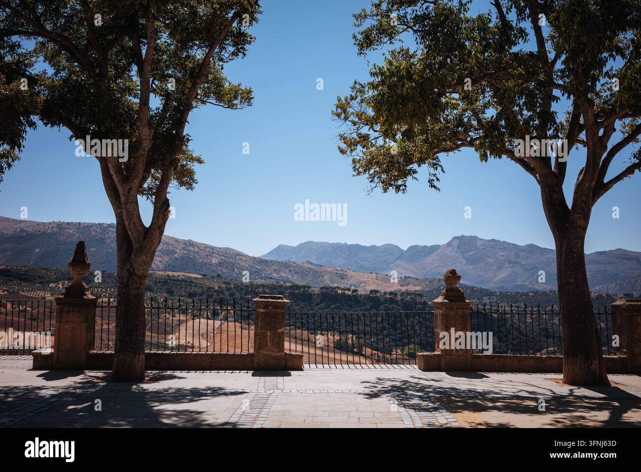 Sonnenbeleuchtete Berglandschaft, gesehen von einer Steinterrasse in Ronda, Spanien, eingerahmt von alten Bäumen, kunstvollen Balustraden und Eisengeländern unter einem klaren Blau Stockfoto