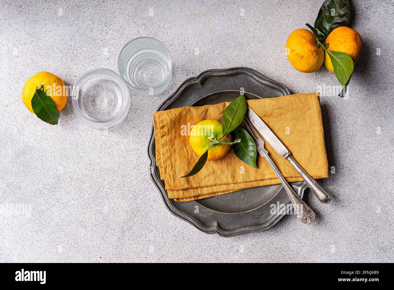 Ansicht von oben eines rustikalen Tischgedecks mit frischen Zitrusfrüchten, grünen Blättern, Glasgeschirr, Besteck, einer Senfserviette und einem Vintage-Metalltablett auf einem Gr Stockfoto