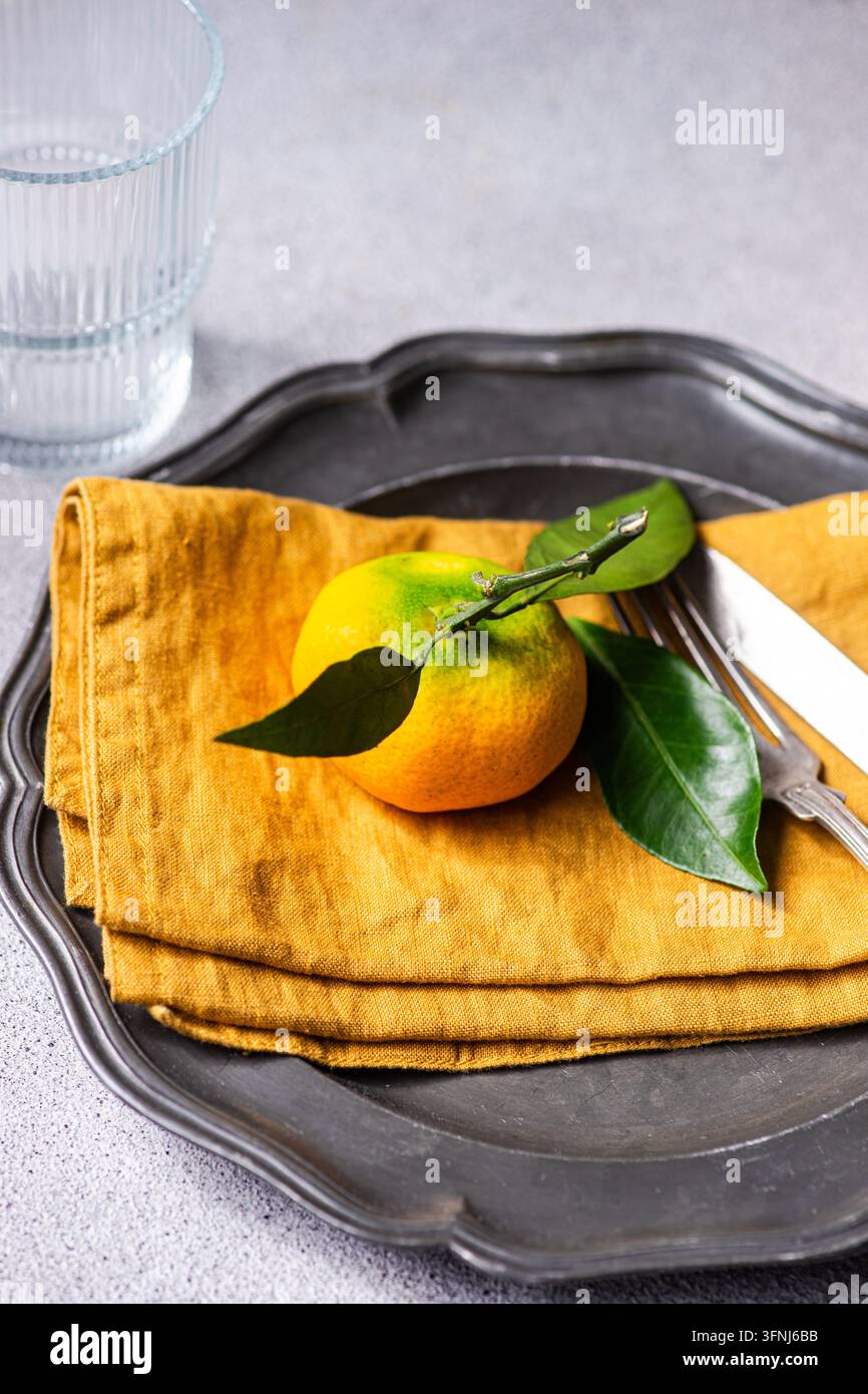 Frische Mandarinen-Zitrusfrüchte mit grünen Blättern liegen auf einem gefalteten Senf-Leinenstoff, arrangiert auf einem dunklen Metalltablett neben einem Glas und silbernem Besteck. Stockfoto