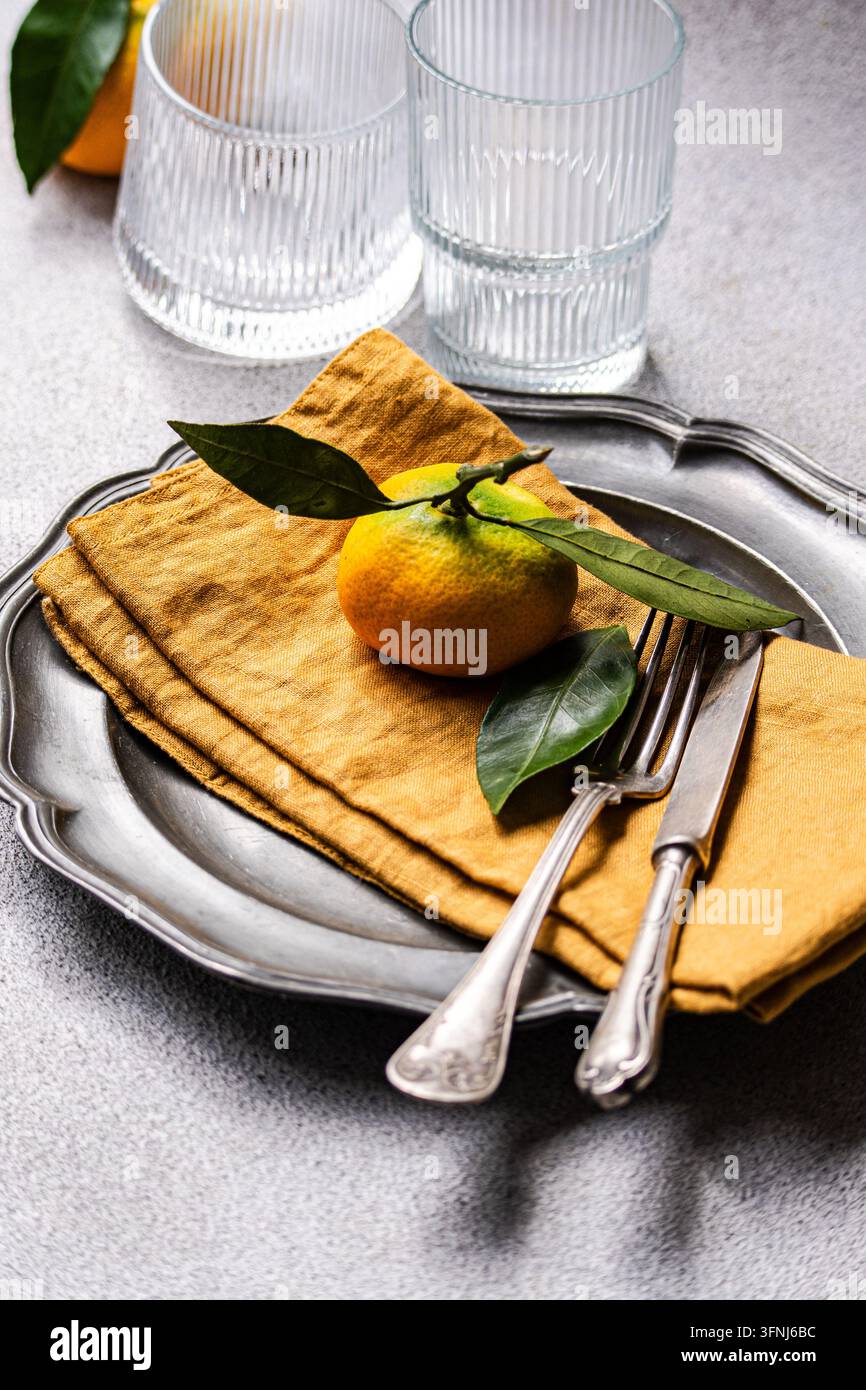 Elegante Tischdekoration mit einer frischen Zitrusfrucht mit grünen Blättern auf goldenen Leinenservietten, arrangiert auf einem Vintage-Silbertray mit Gabel, Messer, an Stockfoto