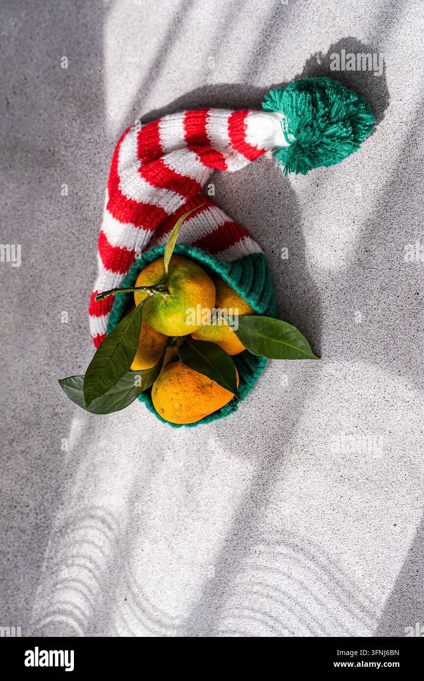 Frische Orangen mit grünen Blättern ruhen in einem rot-weiß gestreiften Weihnachtsstrumpf auf strukturiertem Schnee, beleuchtet von winterlichem Sonnenlicht und dramatischem s Stockfoto