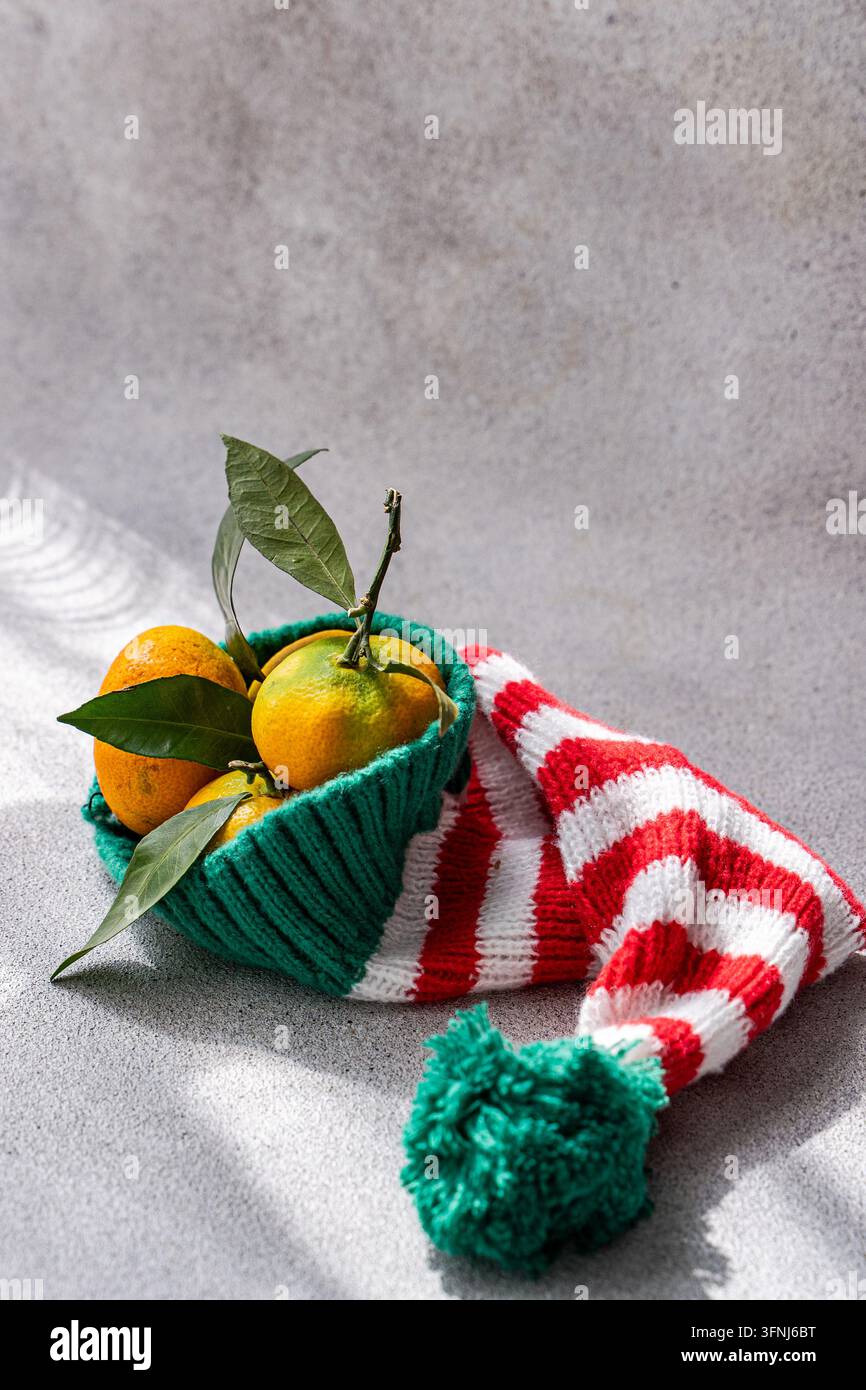 Frische Orangen mit grünen Blättern ruhen in einem rot-weiß-grün gestreiften Weihnachtsstrumpf auf einem grauen strukturierten Hintergrund, was eine festliche Weihnachtsstimmung erzeugt Stockfoto