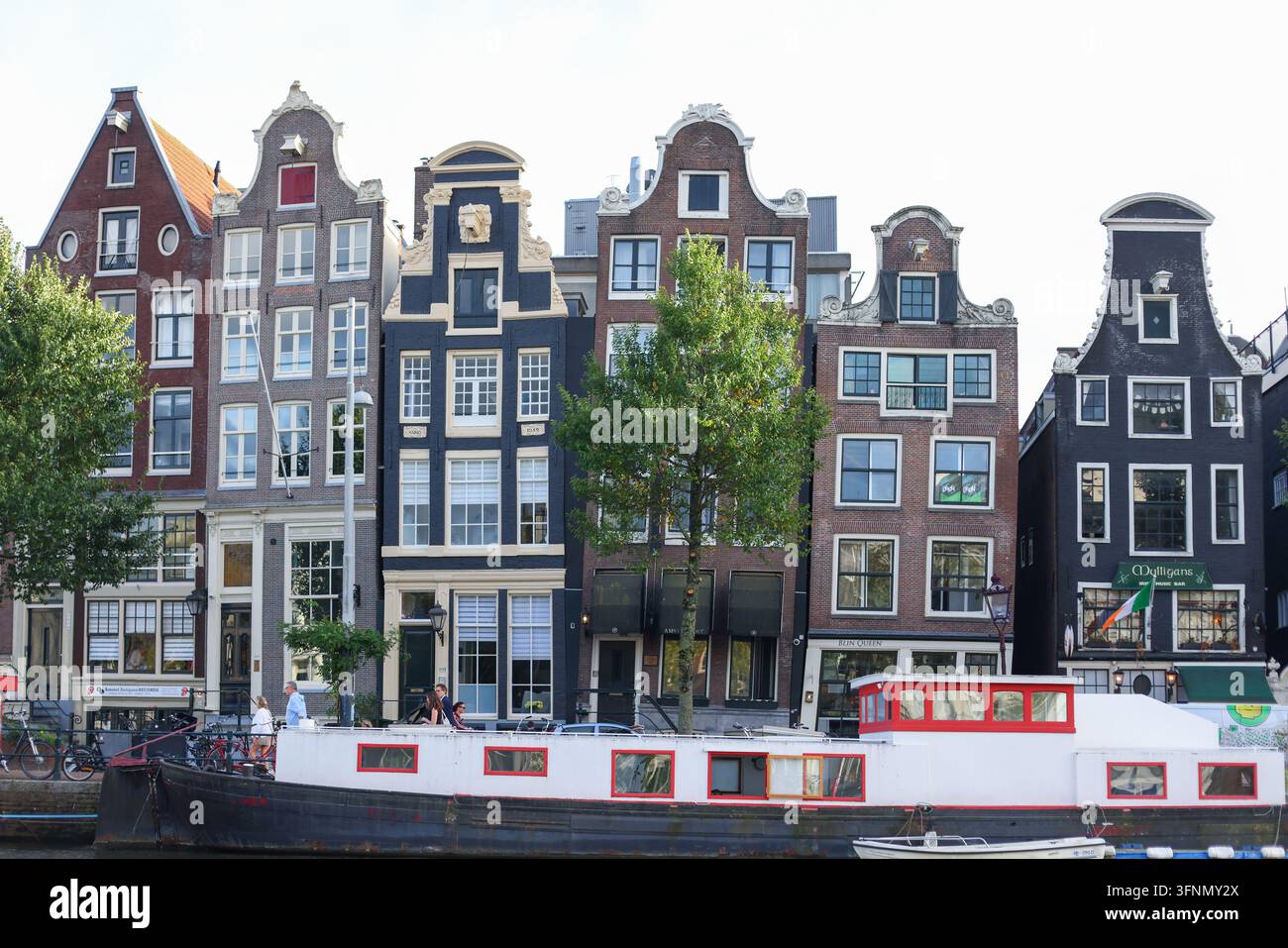 Amsterdam, Niederlande - 1. Oktober 2026: Traditionelle schmale Kanalhäuser und ein vertäutes Hausboot säumen das historische Ufer. Stockfoto