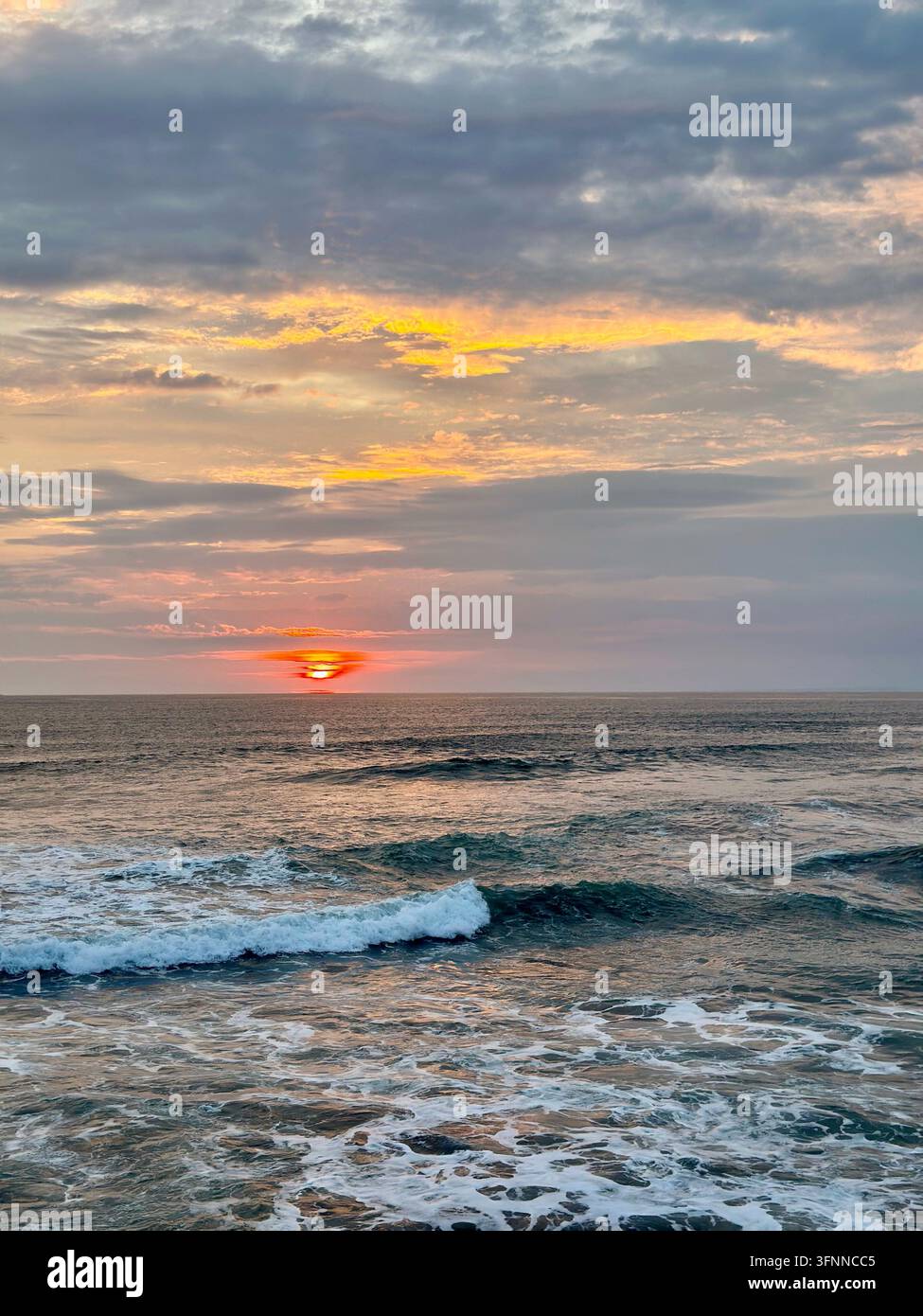 Ein orangefarbener Sonnenuntergang erhellt geschichtete Wolken über brechenden Ozeanwellen und weißem Schaum. - Smartphone-aufgenommenes Stockfoto